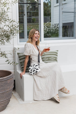 Woman in a white dress sitting on a bench with green cushions, holding a drink, in a bright outdoor setting with black and cream crossbody bag